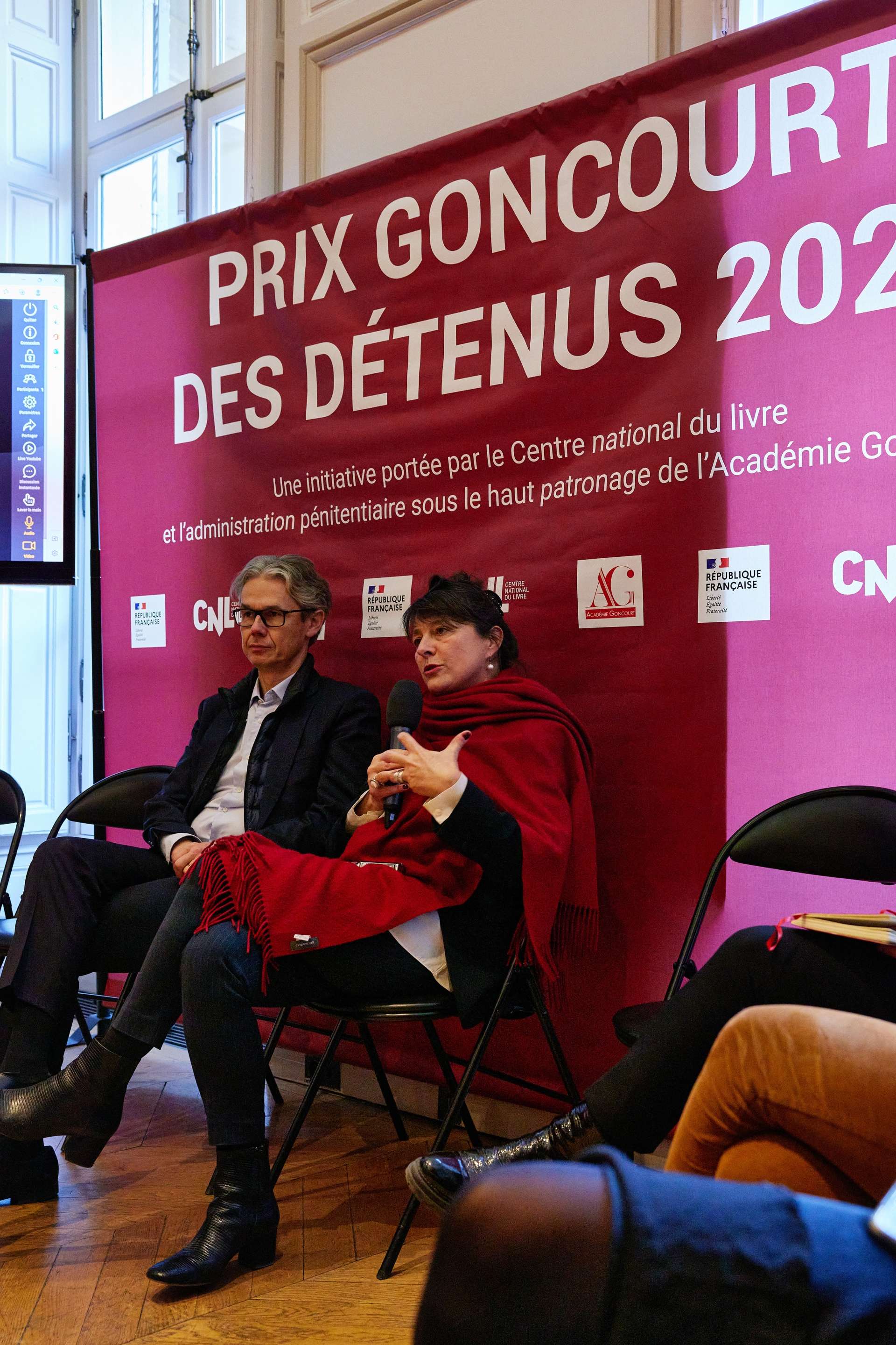 Deux personnes sont assises, l'une d'entre elles tient un micro. Derrière eux, il y a une toile rouge foncée tendue avec l'inscription "Prix Goncourt des détenus 2022"