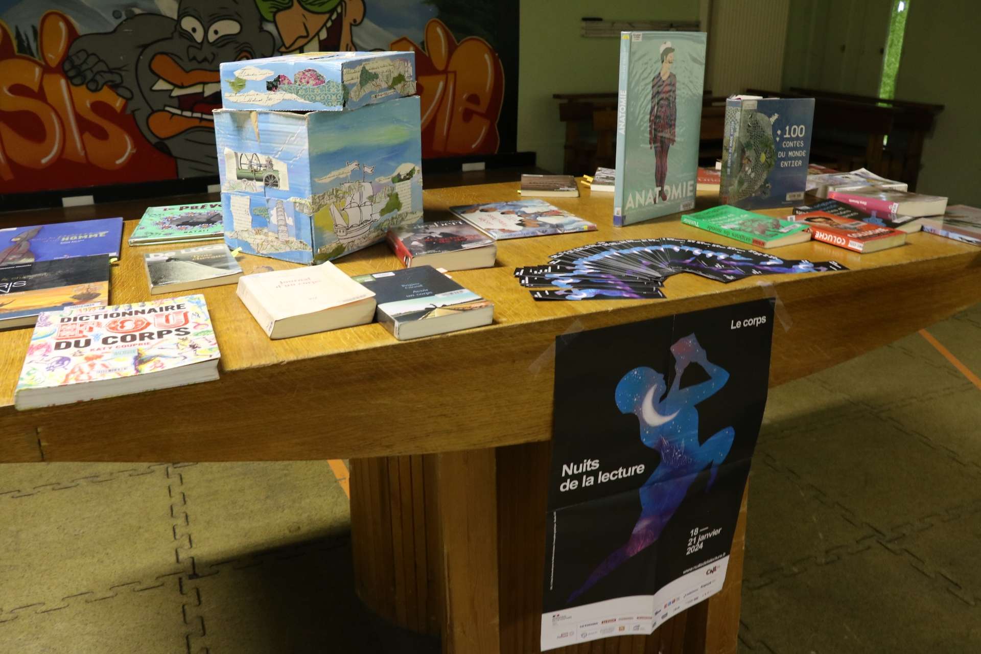 Une vingtaine de livres, avec des formats différents, sont posés à plat sur une table en bois, leurs couvertures bien en évidence. Sur l'un des pieds de la table, une affiche représentant les Nuits de la lecture est collée.