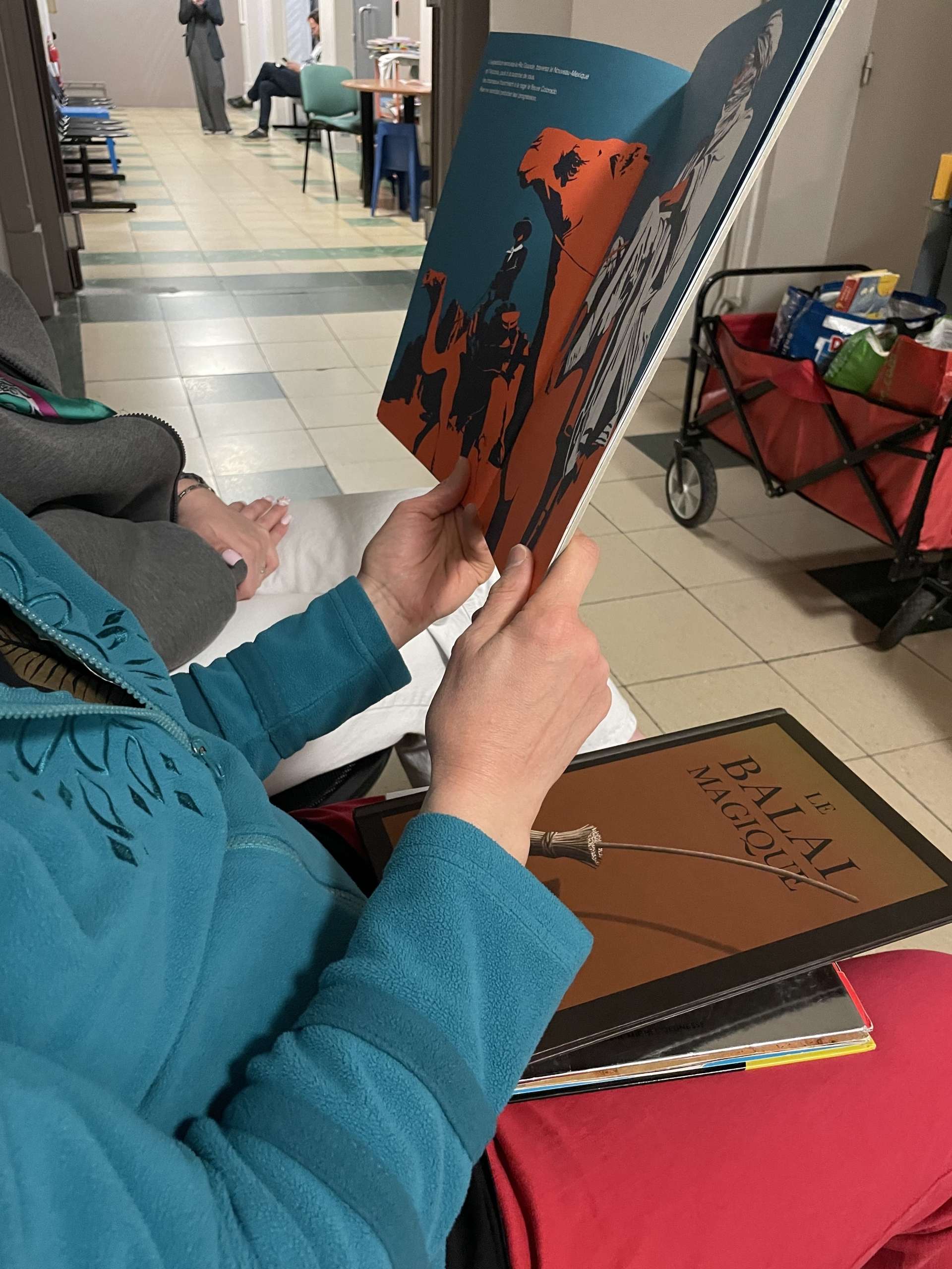 Une personne dont on ne voit pas le visage est assise et tient un livre ouvert dans ses mains. En arrière plan, on peut voir un couloir avec des chaises placées le long des murs.